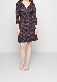 Fioletowa sukienka w kwiatowy wzór, z dekoltem w kształcie litery V, rękawami 3/4, zwężoną talią i rozkloszowanym dołem. Noszona z białymi sandałami na straps.