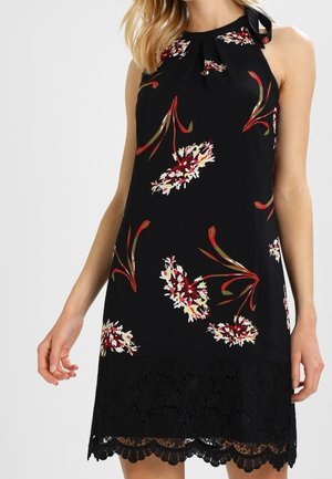 Mouwloze zwarte jurk met rode en witte bloemenprints, halternekstrik en zwarte kanten rand aan de zoom, gedragen door een staande vrouw.