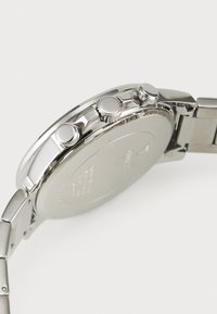 Montre-bracelet en acier inoxydable argenté avec une surface lisse et polie, comportant trois boutons-poussoirs et une couronne texturée, présentant des gravures minimales.