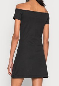 Kvinna klädd i en kort svart avskuren klänning, visad från ryggen mot en neutral ljus bakgrund.