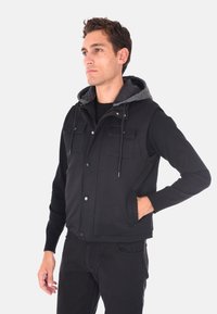 Gilet nero con cappuccio grigio, caratterizzato da un design testurizzato, bottoni a pressione e quattro tasche frontali. Indossato sopra una camicia nera e pantaloni.