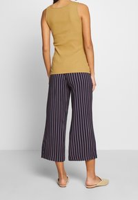 Mujeres vistiendo un top sin mangas mostaza, pantalones anchos recortados en navy con rayas verticales rosas, y zapatos de lona beige, de pie de espaldas.