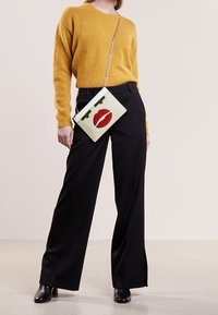 Žlutý fuzzy svetr, černé široké kalhoty a bílá taška s grafickým motivem červených rtů a zelenými akcenty na stříbrném řetízku. Černé boty na podpatku.