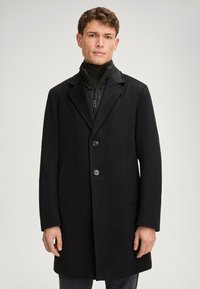 Zwarte jas van een wollen mix met een klassiek ontwerp, twee knopen, gegoten revers en zijzakken, gedragen over een zwarte zip-up jack.