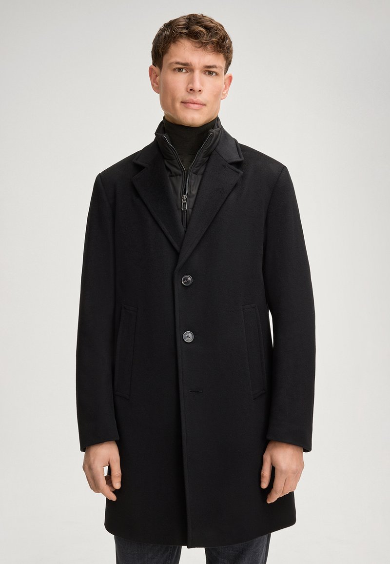Zwarte jas van een wollen mix met een klassiek ontwerp, twee knopen, gegoten revers en zijzakken, gedragen over een zwarte zip-up jack.