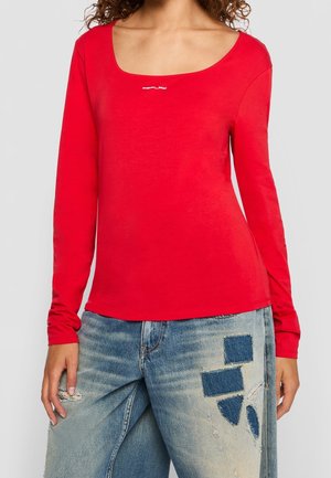 Kvinde iført en rød langærmet bluse med rund halsudskæring og lille hvidt logo, kombineret med lyseblå slidte jeans med mørkeblå lapper.
