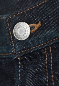 Κοντινή λήψη σκούρου μπλε τζιν υφάσματος με πορτοκαλί ραφές και μεταλλικό κουμπί με το χαραγμένο επανειλημμένα τη λέξη "WEEKDAY".