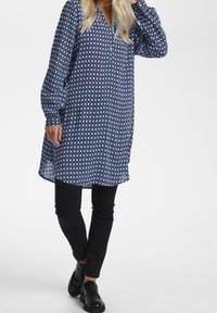 Niebieska tunika w wzory z długimi rękawami, z zapięciem na guziki z przodu i luźnym krojem. Noszona na czarnych spodniach i czarnych butach.