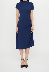 Mörkblå klänning med korta ärmar, figurnära överdel och A-linjeformad kjol. Slät tyg med rynkdetalj på sidan, bärs med svarta klackar.