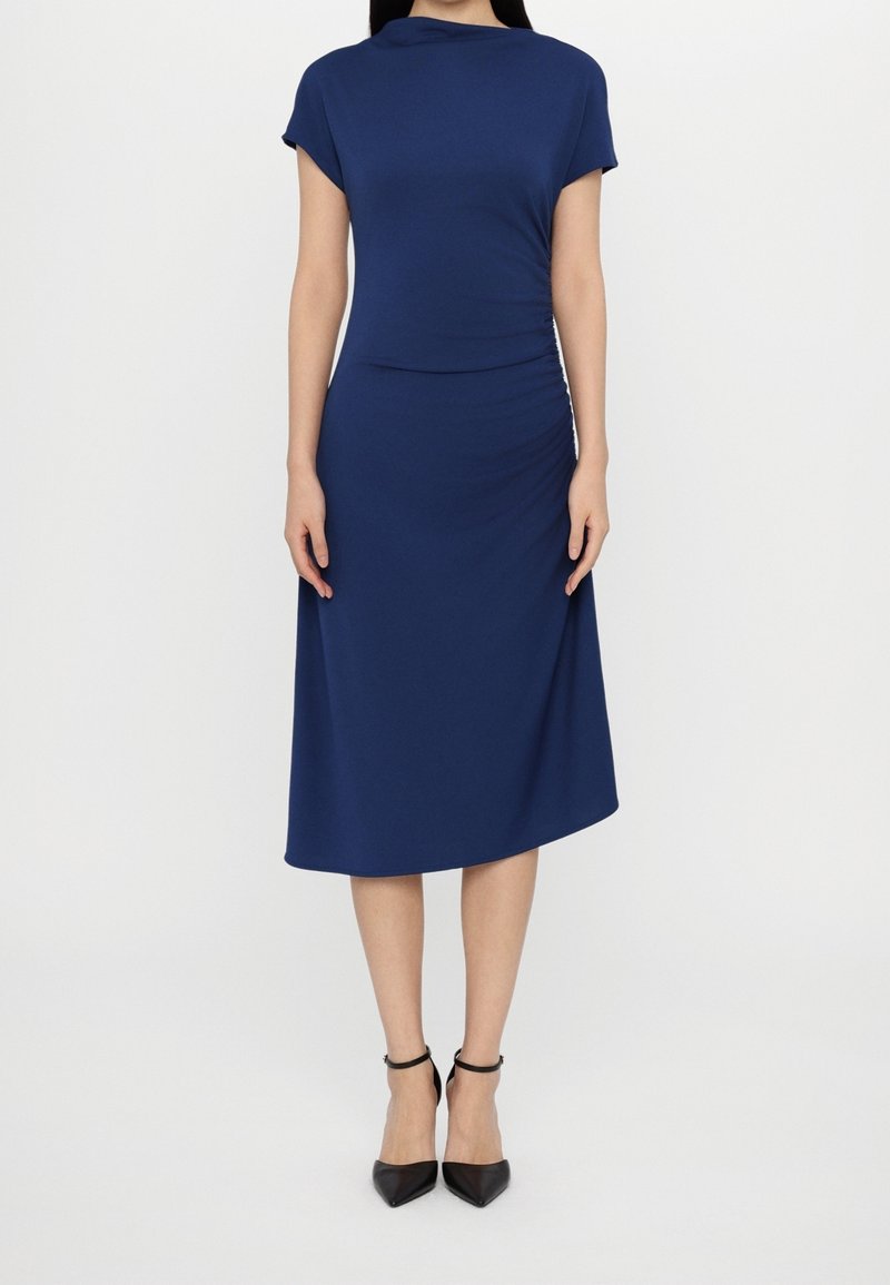 Mörkblå klänning med korta ärmar, figurnära överdel och A-linjeformad kjol. Slät tyg med rynkdetalj på sidan, bärs med svarta klackar.