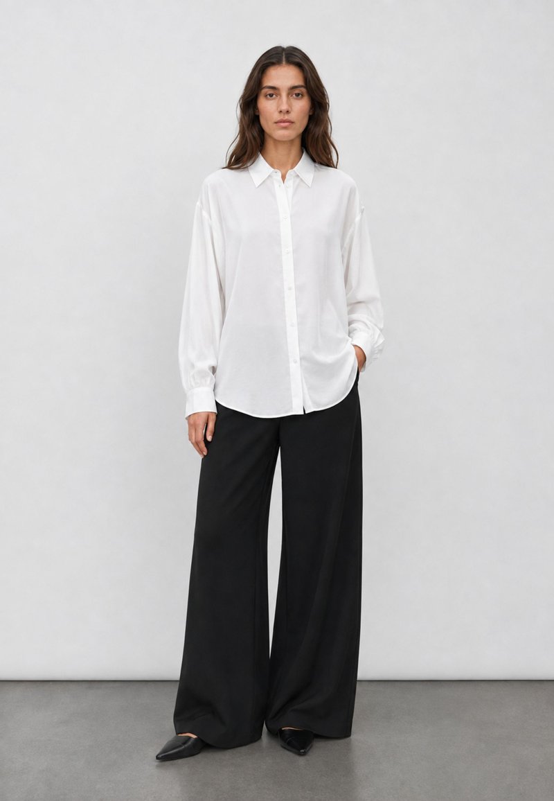 Femme portant une chemise blanche ample boutonnée, un pantalon large noir et des chaussures noires pointues, debout devant un fond gris clair uni.