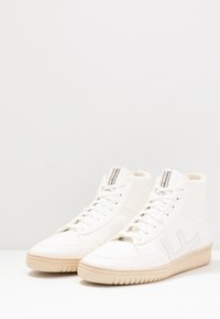Białe sneakersy typu high-top wykonane z gładkiej skóry z teksturowaną podeszwą, z sznurowadłami i akcentem logo po boku.