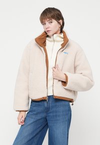 Persoană tânără îmbrăcată cu o jachetă de fleece bej cu margini maro, un pulover alb cu fermoar pe jumătate și blugi largi albaștri, stând pe un fundal simplu.