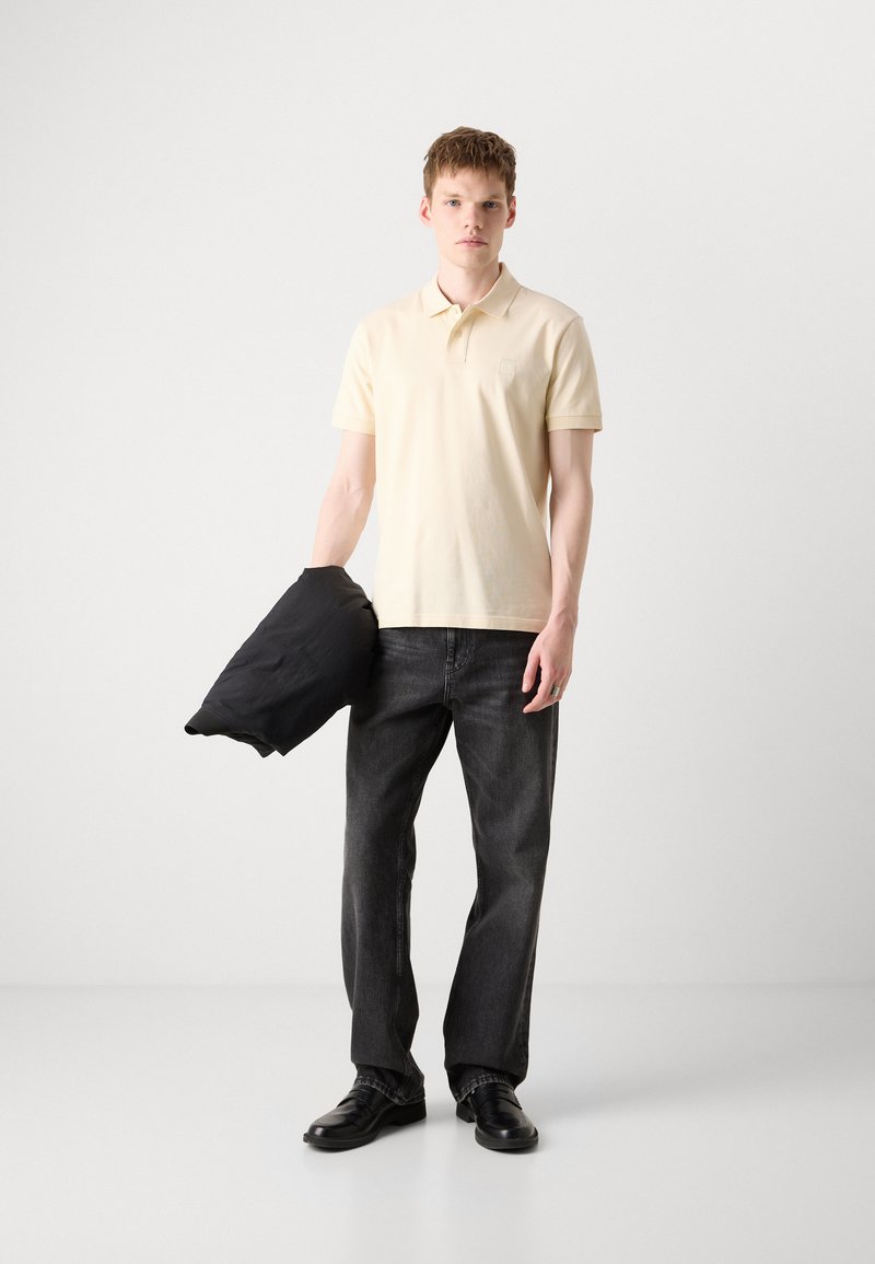 Beige Poloshirt, dunkelgraue Jeans und schwarze Schuhe. Das Model hält eine schwarze Jacke. Einfaches Design, glatte Textur und lockere Passform.