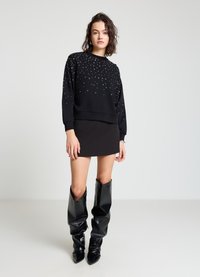 Pull-over noir avec des strass éparpillés, col rond et ourlet côtelé, accompagné d'une mini-jupe noire et de bottes montantes brillantes jusqu'aux genoux.