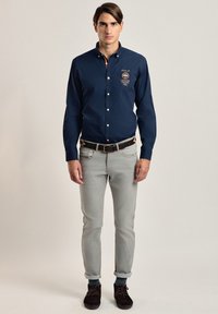 Camisa de botones azul marino con collar, detalles de logotipo y parche; combinada con jeans gris claro y zapatos oscuros, sobre un fondo neutro.