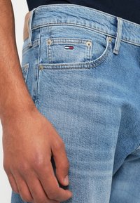 Ανοιχτό μπλε τζιν με μια διακριτική υφή φθοράς; διαθέτει ένα μικρό κεντημένο λογότυπο σε κόκκινο, άσπρο και μπλε στην πίσω τσέπη.