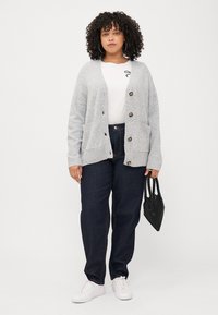 A woman with curly hair stands confidently, wearing a gray cardigan over a white shirt and dark jeans, holding a black beaded bag.