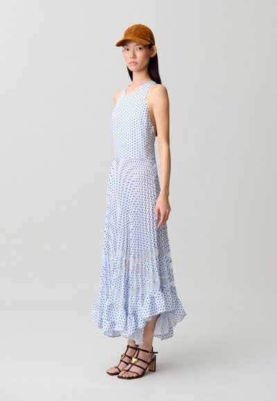 Donna che indossa un vestito azzurro chiaro senza maniche a pois con orlo arricciato, cappello marrone e sandali neri con cinturini e dettagli a forma di cuore dorati.