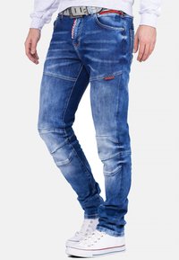 Jean en denim bleu avec une coupe slim, présentant des zones usées, des détails de couture et des accents rouges contrastants. Associés à des baskets blanches.