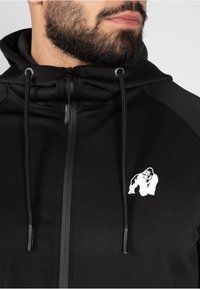 Felpa nera con zip, dal tocco morbido, con un logo di gorilla bianco sul lato sinistro del petto e con lacci neri regolabili sul cappuccio.