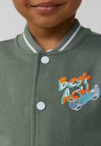 Grüne Baumwolljacke mit einem gerippten Kragen, weißen Streifen und Druckknöpfen. Verfügt über bunten gestickten Text und ein Skateboard-Graphic.