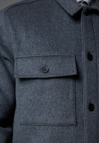 Grå uldjakke med en knaplukket brystlomme og sorte knapper. Stoffet er blødt med synlige vævemønstre.