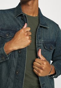 Jeansjacke mit dunkler Waschung, ausgestattet mit Metallknöpfen, Klappentaschen und einem klassischen Kragen. Getragen über einem einfarbigen olivgrünen T-Shirt.