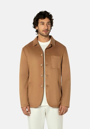 Man met krullend haar, gekleed in een bruin jasje met knopen, gedragen over een wit shirt en witte broek, staand en iets naar rechts gericht.