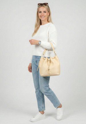 Hymytteleva nainen valkoisessa villapaidassa, vaaleansinisissä farkuissa, valkoisissa lenkkareissa ja päässään aurinkolasit pitää beigenväristä nahkalaukkua käsivarrellaan.