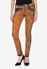 Jeans en denim orange avec des motifs de éclaboussures bleues et noires. Design ajusté, avec une ceinture cloutée et des passants de ceinture. Des talons hauts complètent le look.