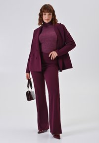 Bordeaux pak met een getailleerde blazer en uitlopende broekspijpen, gecombineerd met een sprankelende coltrui. Accessoires zijn een kleine handtas en puntige schoenen.