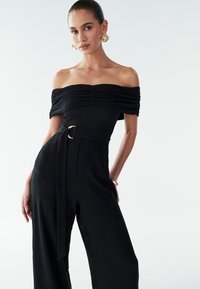 Sort off-shoulder jumpsuit i tekstureret stof med rynket overdel, vid ben-design og talje med bælte, der har en guldfarvet rund detalje.
