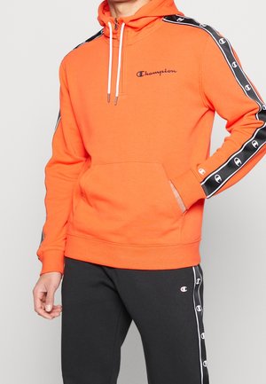 Orange sweatshirt med hætte, hvide snore og forlomme. Har sorte sidelommer med logo-accenter. Kombineret med sorte bukser.