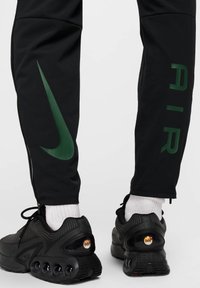 Pantalons de sport noirs avec un logo Nike vert et le texte "AIR". Le design comprend un ourlet zippé et est associé à des baskets noires et des chaussettes blanches.