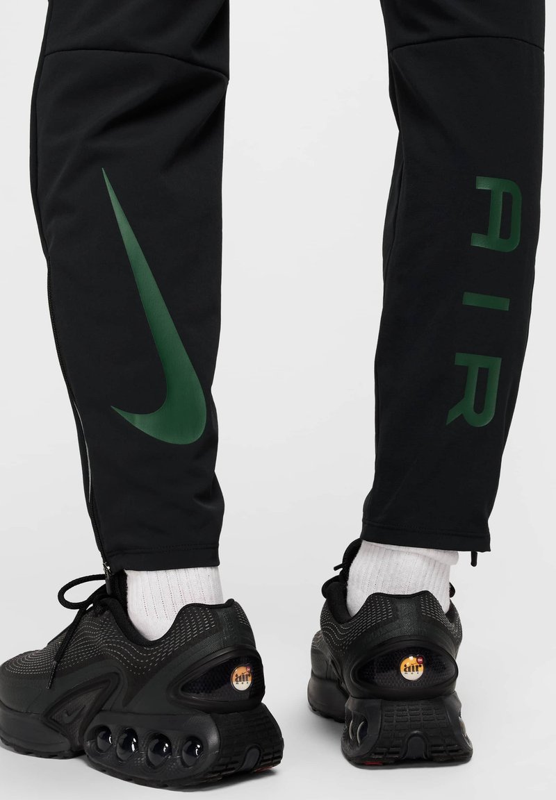 Pantalons de sport noirs avec un logo Nike vert et le texte "AIR". Le design comprend un ourlet zippé et est associé à des baskets noires et des chaussettes blanches.