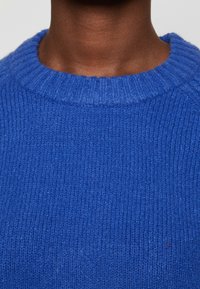 Pull bleu tricoté avec un col rond côtelé. Le tissu semble doux et texturé, présentant des lignes verticales dues au motif du tricot.
