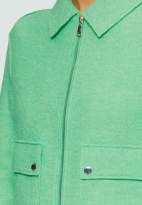 Veste verte texturée avec un zip sur le devant, un col pointu et deux poches avec bouton. Présente des accents en matériel argenté.