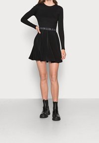 Black long-sleeve dress with a fitted bodice and flared skirt. Features a Calvin Klein Jeans logo waistband and smooth fabric texture.