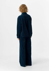 Person mit lockigem blonden Haar, der ein dunkelblaues Cordhemd mit langen Ärmeln und weiten Hosen trägt, steht mit dem Rücken zu einer einfarbigen Hintergrund.