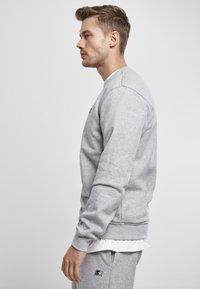 Sudadera gris hecha de mezcla de algodón, con puños y dobladillo acanalados, y un pequeño logo en el pecho. El modelo se muestra de perfil.