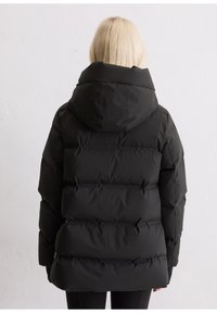 Eine blonde Person trägt eine schwarze, gepolsterte Winterjacke mit einer großen Kapuze und steht mit dem Rücken zu einem schlichten weißen Hintergrund.