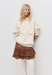 Felpa oversize color crema con tasca frontale e ricamo rosso, abbinata a una gonna strati multicolore e testurizzata.