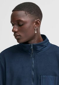 Giacca in pile blu navy con colletto alto, chiusura con zip e tasca sul petto. Presenta una fodera interna liscia e dettagli con zip sulla parte anteriore.