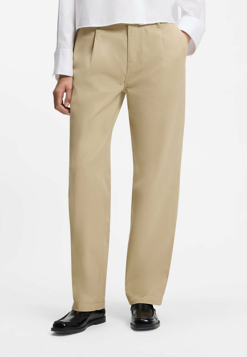 Pantalon beige sur mesure avec plis devant porté avec des mocassins noirs et une chemise blanche à manches longues, mains dans les poches, sur un fond uni.