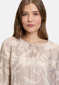 Lekka, przejrzysta bluzka w miękkim beżu z kwiatowym wzorem. Posiada zebrany dekolt i płynny krój.