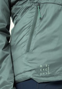 Grüne Jacke mit schräger Reißverschlusstasche und dem Schriftzug "Recycled Inside" nahe dem Saum, getragen über dunkelblauer Hose.