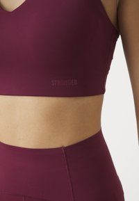 Bordo sportski grudnjak od glatkog materijala, s V-izrezom i diskretnim logom. Glatka tekstura s elegantnim krojem.