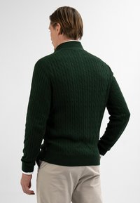 Man gezien van achteren in een donkergroene kabeltrui en lichtbeige broek, staande tegen een effen witte achtergrond.