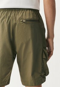 Shorts vert olive en tissu léger, avec une taille élastique, une poche zippée et une poche latérale de type cargo. Fini lisse et texturé.